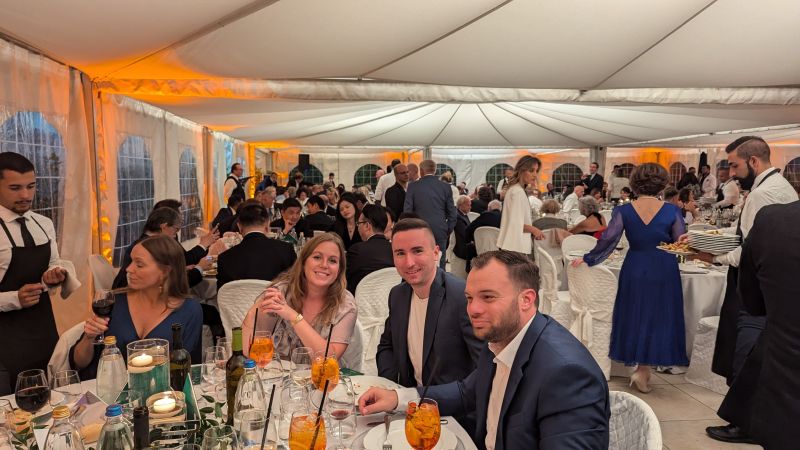 Festliche Abendveranstaltung in einem großen, beleuchteten Zelt. Im Vordergrund sitzen mehrere Gäste in eleganter Kleidung an einem gedeckten Tisch, während im Hintergrund zahlreiche weitere Gäste speisen und Servicepersonal Essen serviert.